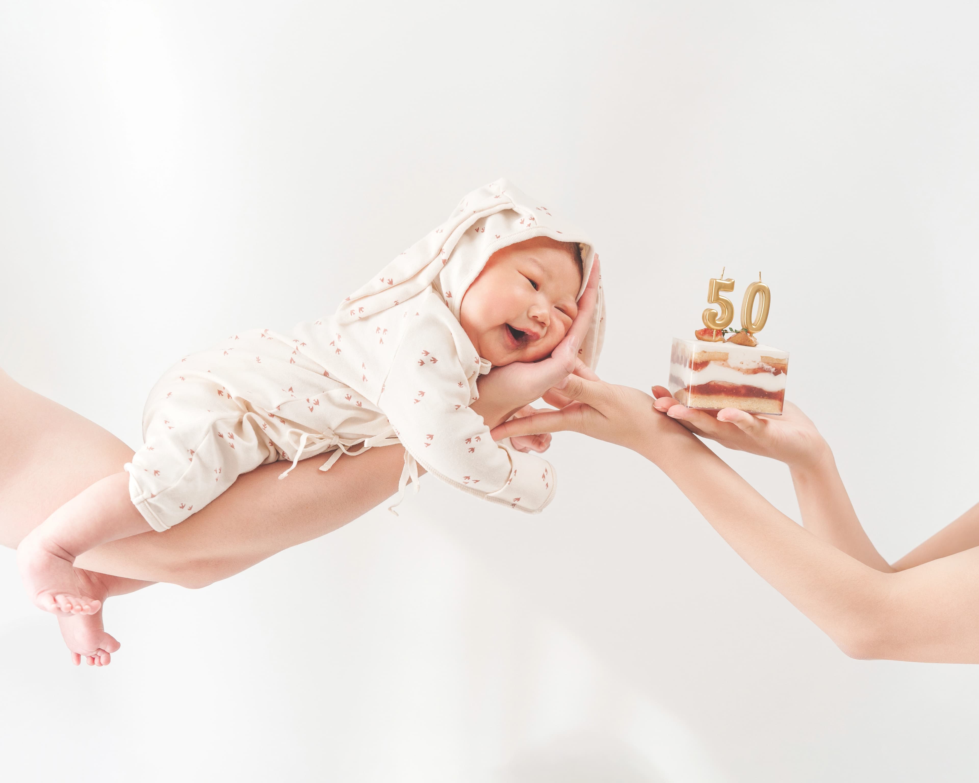 하얀 배경 앞에서 꽃무늬 옷을 입은 아기가 케이크 위에 꽂힌 숫자 50 초를 향해 손을 뻗으며 밝게 웃고 있는 모습.