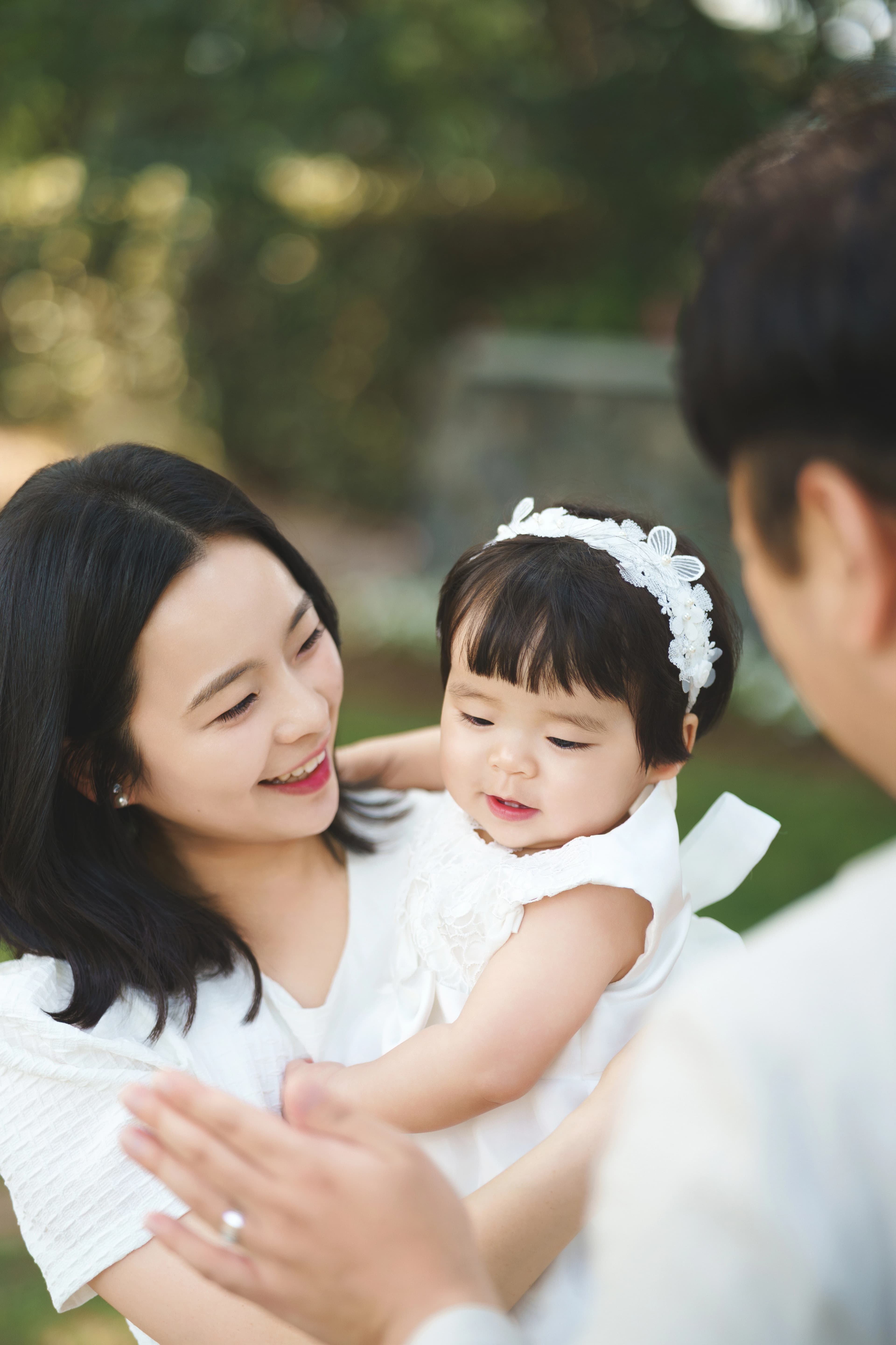 따뜻한 햇살 아래 야외에서 흰색 옷을 맞춰 입은 젊은 부부가 아기를 품에 안고 다정하게 미소 지으며 행복한 시간을 보내는 모습.