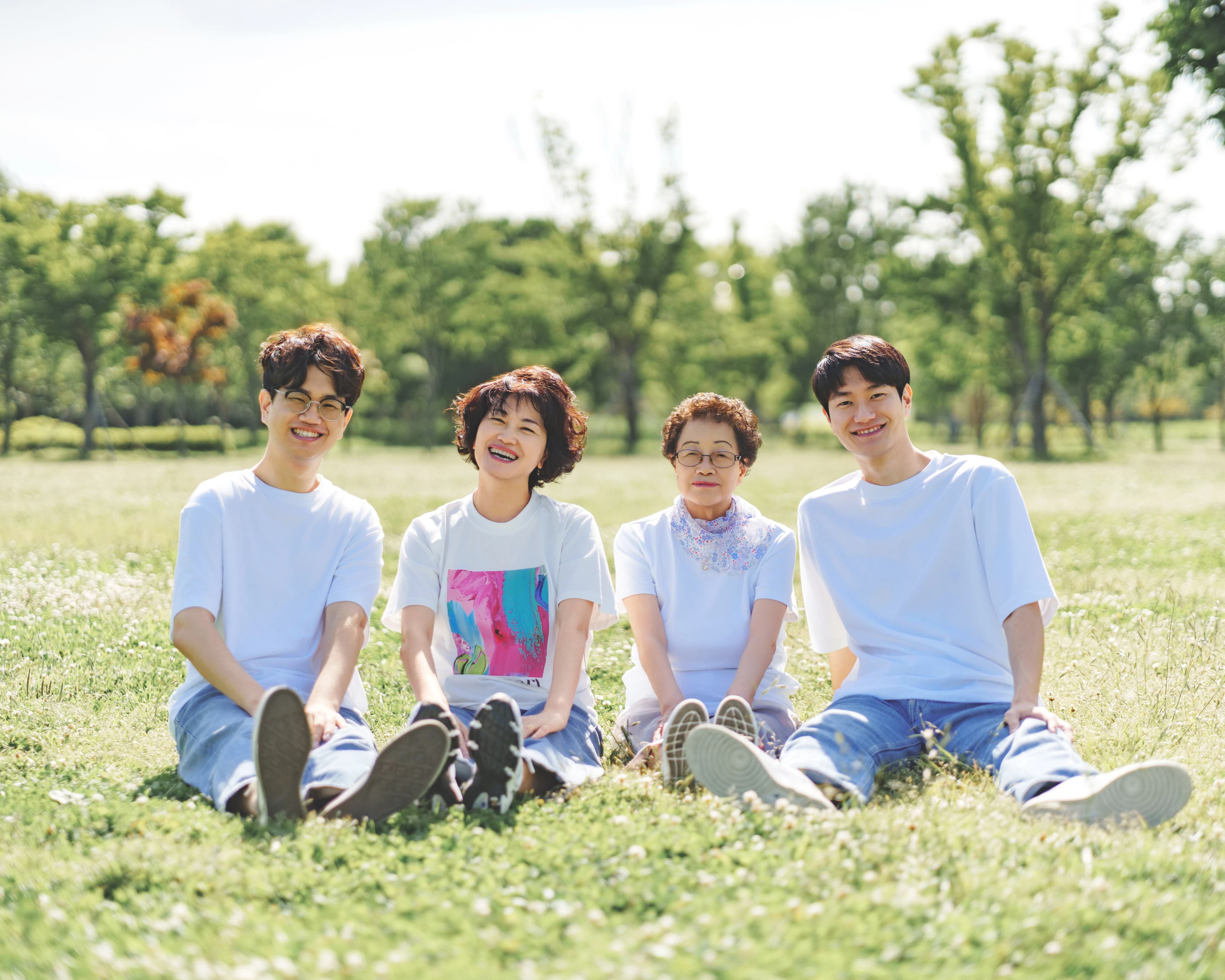 푸른 잔디밭 위에서 네 명의 사람들이 밝게 웃으며 나란히 앉아 여유로운 시간을 보내고 있는 모습.