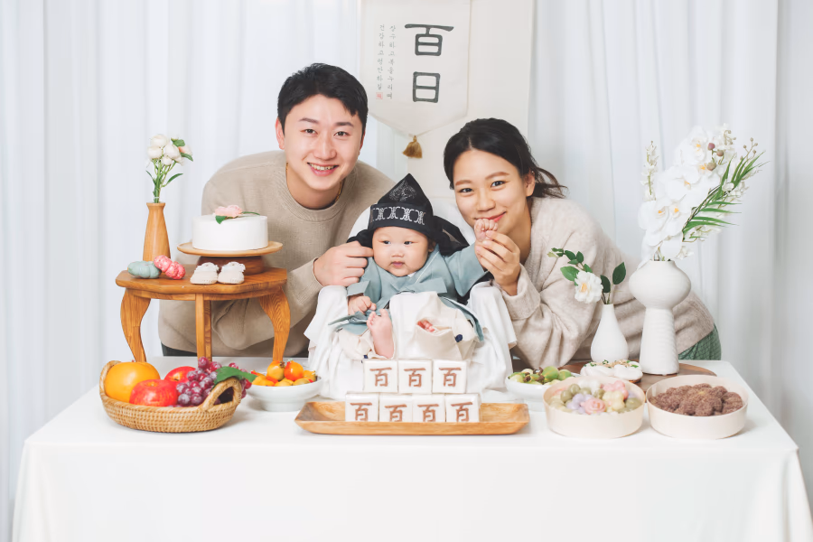 백일잔치 상 앞에 앉아 전통 의상을 입은 아기를 가운데 두고 밝게 웃고 있는 젊은 부부의 행복한 가족사진.