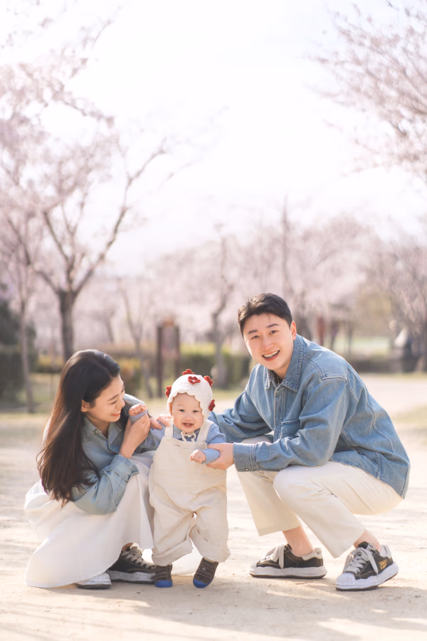 벚꽃이 만개한 공원에서 데님 재킷을 맞춰 입은 젊은 부부가 귀여운 아기와 함께 밝게 웃으며 야외 나들이를 즐기는 행복한 모습.
