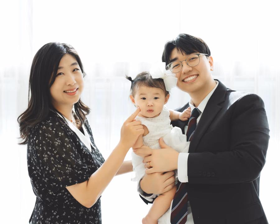 검은색 정장을 입은 남성과 꽃무늬 원피스를 입은 여성이 밝은 실내에서 아기를 품에 안고 다정하게 미소 지으며 포즈를 취하고 있는 모습.