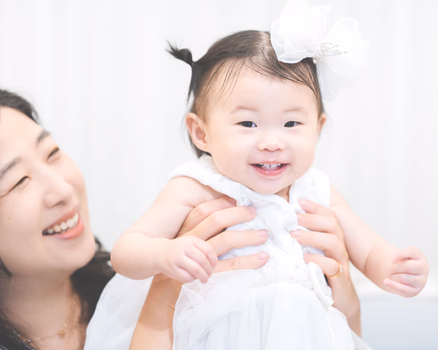 하얀 드레스와 꽃 장식을 한 아기가 밝게 웃고 있고, 옆에서 여성이 다정하게 바라보며 미소 짓는 따뜻한 분위기의 사진.
