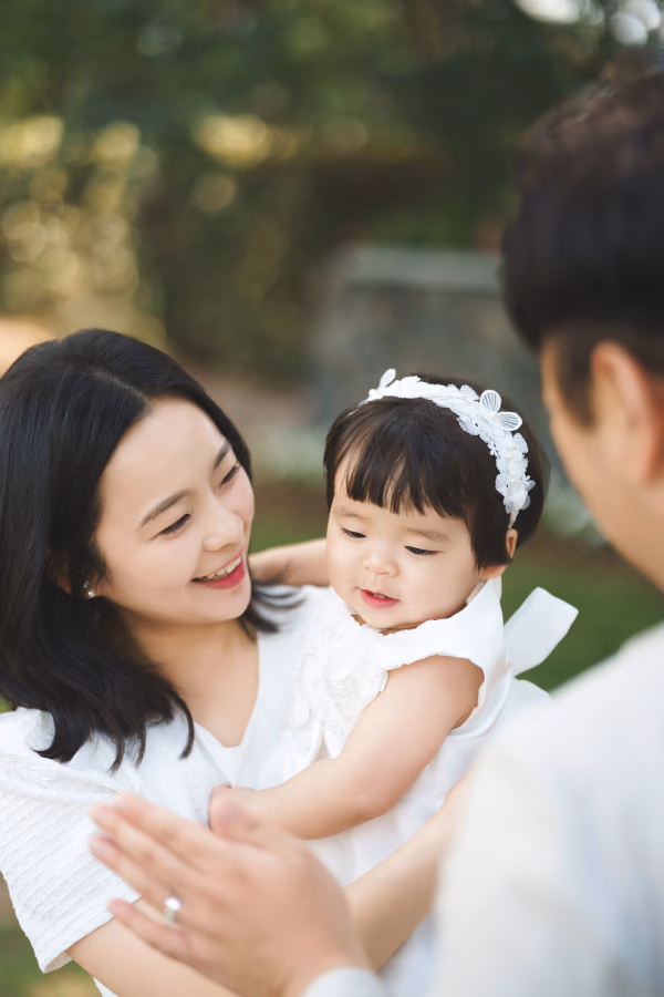 따뜻한 햇살 아래 야외에서 흰색 옷을 맞춰 입은 젊은 부부가 아기를 품에 안고 다정하게 미소 지으며 행복한 시간을 보내는 모습.