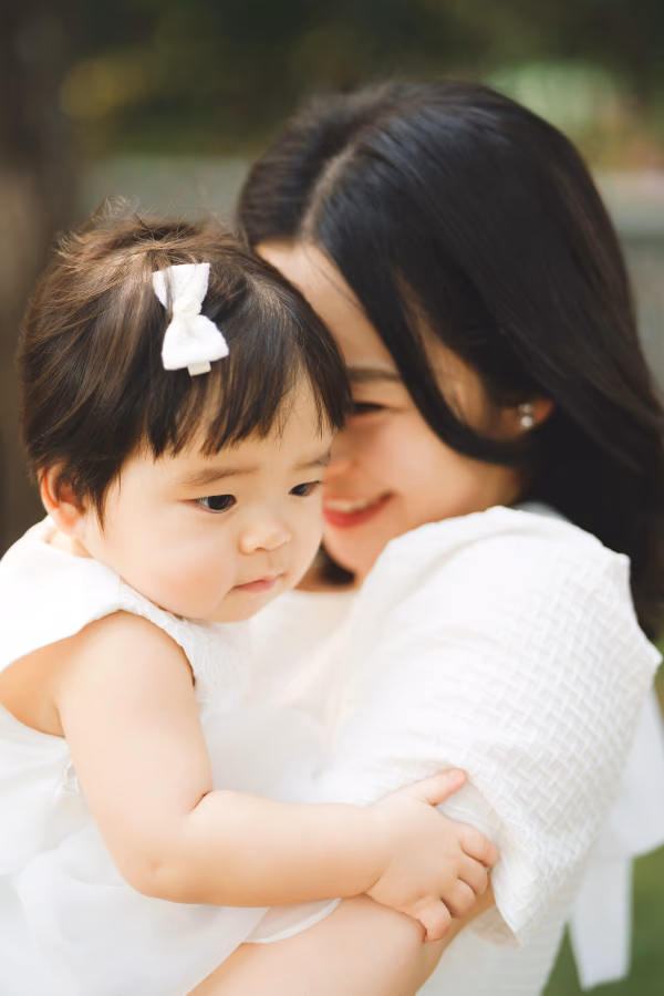따뜻한 햇살 아래 하얀 옷을 입은 아기를 품에 안고 미소 짓고 있는 엄마의 다정한 모습.