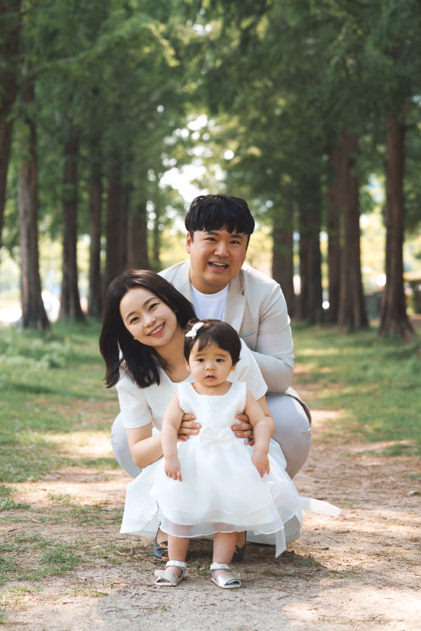 푸른 나무들이 늘어선 공원에서 흰색 옷을 맞춰 입은 행복한 가족이 어린 딸과 함께 다정하게 포즈를 취하고 있는 모습.