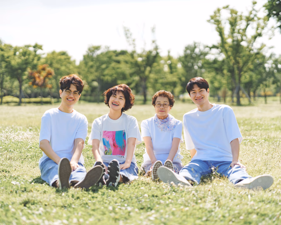 푸른 잔디밭 위에서 네 명의 사람들이 밝게 웃으며 나란히 앉아 여유로운 시간을 보내고 있는 모습.