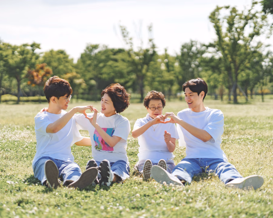 푸른 잔디밭 위에서 흰 티셔츠를 맞춰 입은 네 명의 가족이 서로 손으로 하트 모양을 만들며 밝게 웃고 있는 화목한 모습.