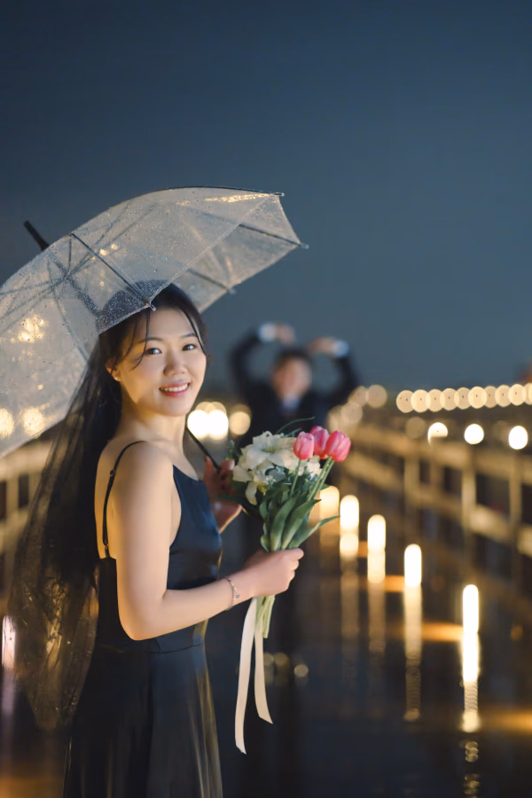 투명한 우산을 쓰고 꽃다발을 든 여성이 밤의 은은한 조명이 빛나는 야외에서 미소 짓고 있는 로맨틱한 모습.