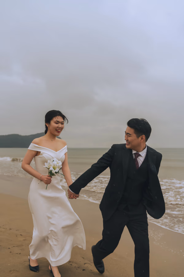 해변에서 웨딩드레스와 수트를 차려입은 신랑 신부가 서로 손을 잡고 즐겁게 뛰어노는 로맨틱한 순간.