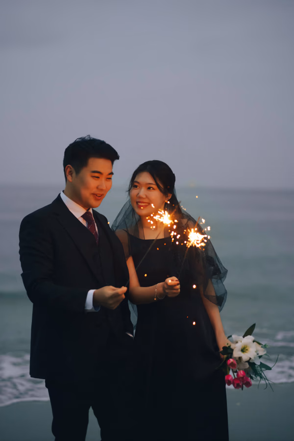 해질녘 바닷가에서 검은색 드레스와 수트를 입은 커플이 스파클러를 들고 행복하게 미소 짓는 로맨틱한 모습.