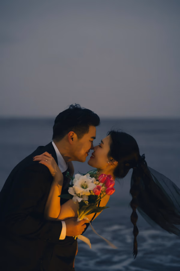해질녘 바닷가를 배경으로 정장을 입은 신랑과 웨딩드레스를 입은 신부가 서로를 다정하게 바라보며 포옹하고 있는 로맨틱한 웨딩 사진.